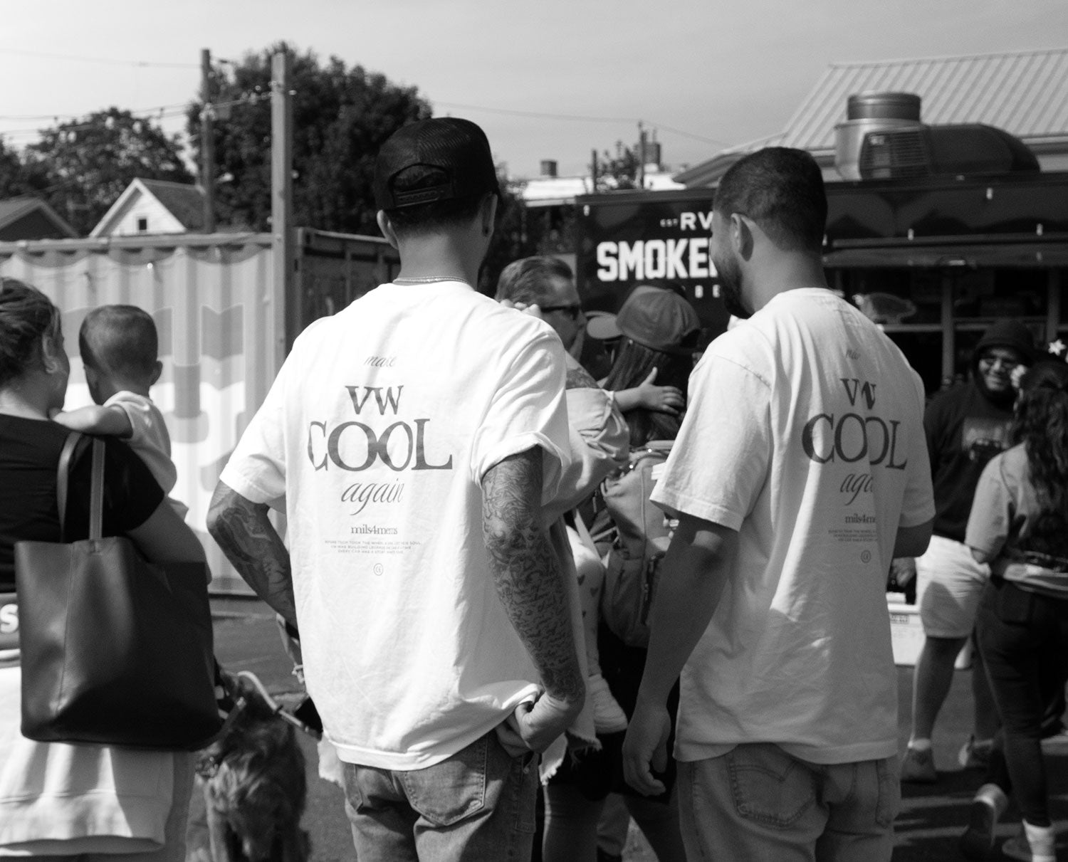 Black and white photo of two Volkswagen enthusiasts wearing 'Make VW COOL Again' t-shirts standing in a crowd at a car show, with a food truck in the background.